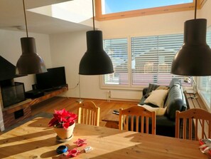 Eine moderne Küche mit Pendelleuchten, einem Esstisch mit Stühlen, einem Fernseher und einem großen Fenster mit Jalousien.