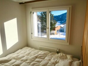 Ein Schlafzimmer mit einem Bett, einem Fenster mit Blick auf eine verschneite Landschaft und Berge sowie einer Heizung.