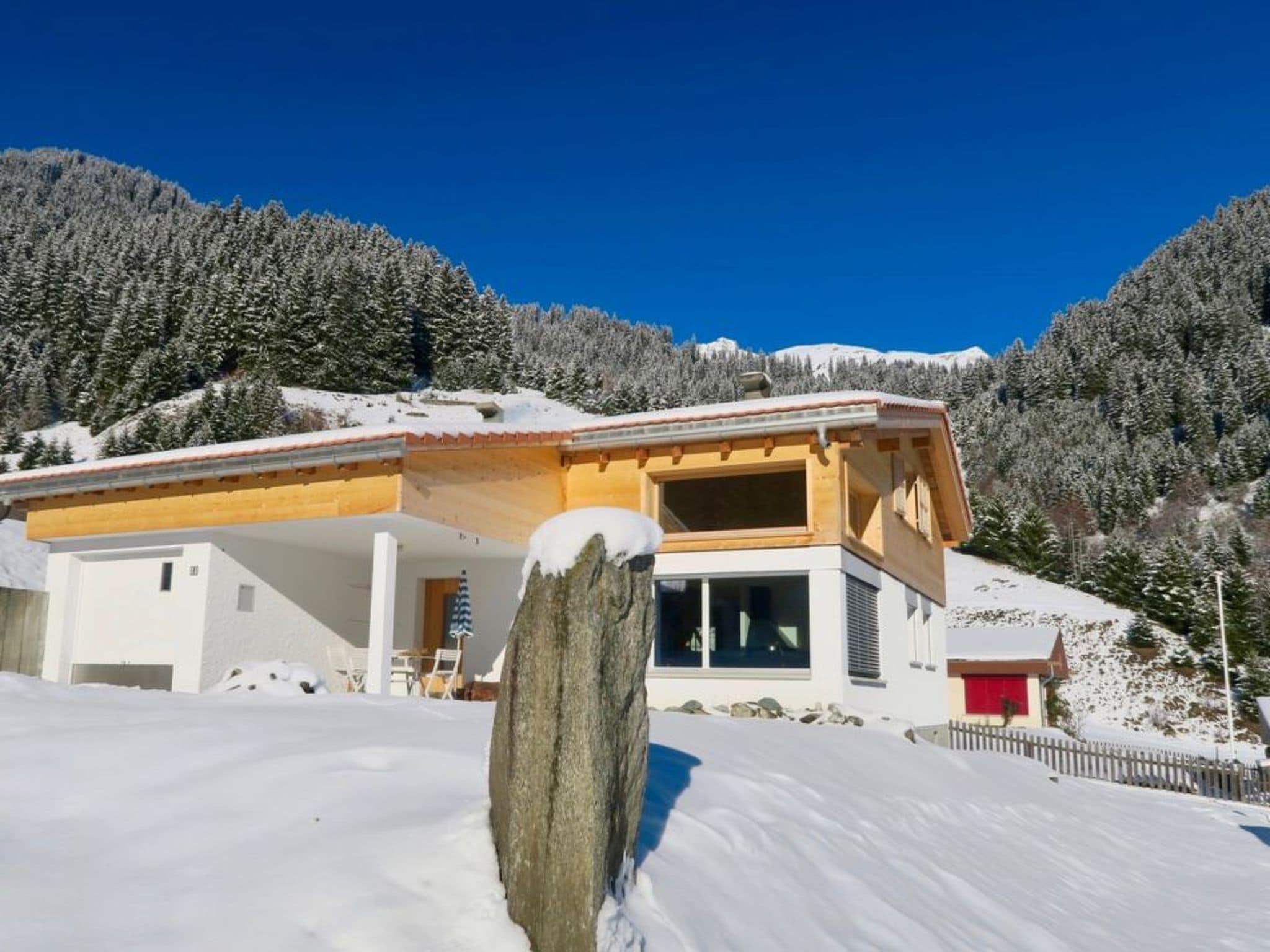 A modern chalet with a large porch, a red garage, and a snowy mountain backdrop.