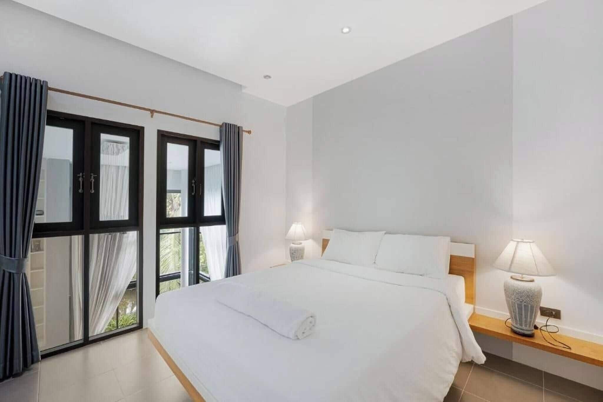 A bedroom with a bed, bedside lamps, a window with curtains, and a view of greenery outside.