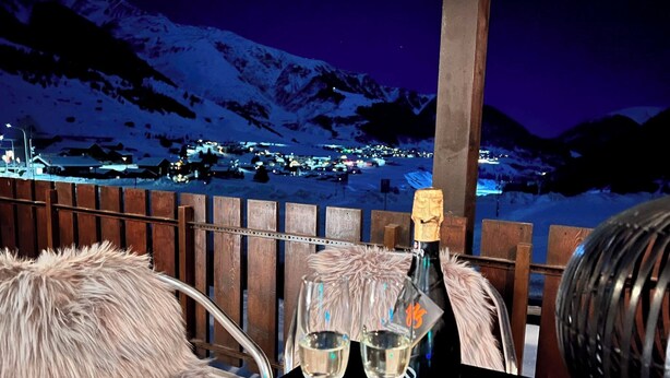 Een tafel met een champagnefles en twee glazen, een fauteuil bekleed met bont, en een uitzicht op een besneeuwde stad 's nachts.