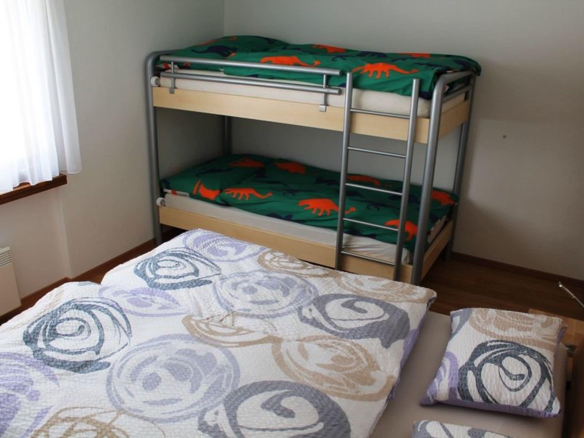 A bunk bed with a green dinosaur-themed mattress cover, a lower bunk with a patterned blanket, and a single bed with a swirl-patterned blanket.