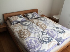 A double bed with a patterned bedspread and two pillows, a bedside table, and a chair. - Tgèsa Palits Hagenbuch, (Sedrun), 107.01 by Interhome (Sedrun)
