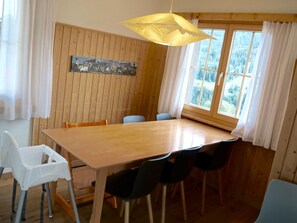 A dining area with a wooden table, chairs, a hanging light fixture, and a window with curtains. - Ferienhaus Kuster, (Sedrun), 006.01 by Interhome (Sedrun)
