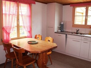 Un coin repas avec une table et des chaises en bois, une cuisine équipée d’un évier et de placards, et une fenêtre agrémentée de rideaux à carreaux.