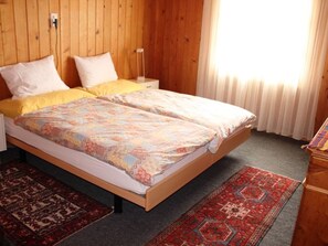 Ein Doppelbett mit blumigem Überwurf, zwei weiße Kissen, ein hölzerner Kopfteil, ein Nachttisch, ein Teppich mit roten und blauen Mustern und eine Holzkombination.