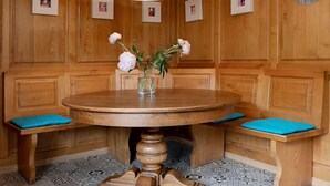 Une pièce aux murs lambrés, avec une table ronde en bois, des bancs dotés de coussins bleus, un luminaire blanc suspendu au plafond et une horloge au mur.