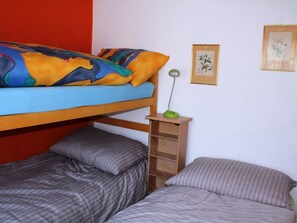 Uma cama de beliche com um cobertor e almofadas coloridos, uma mesinha de cabeceira com abajur, e duas fotos emolduradas na parede.