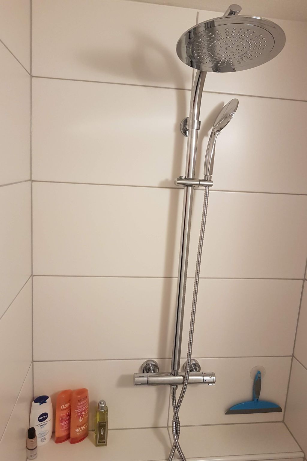 Una doccia con soffione a mano e soffione fisso, uno spazzolino esfoliante e articoli da toilette sul pavimento.