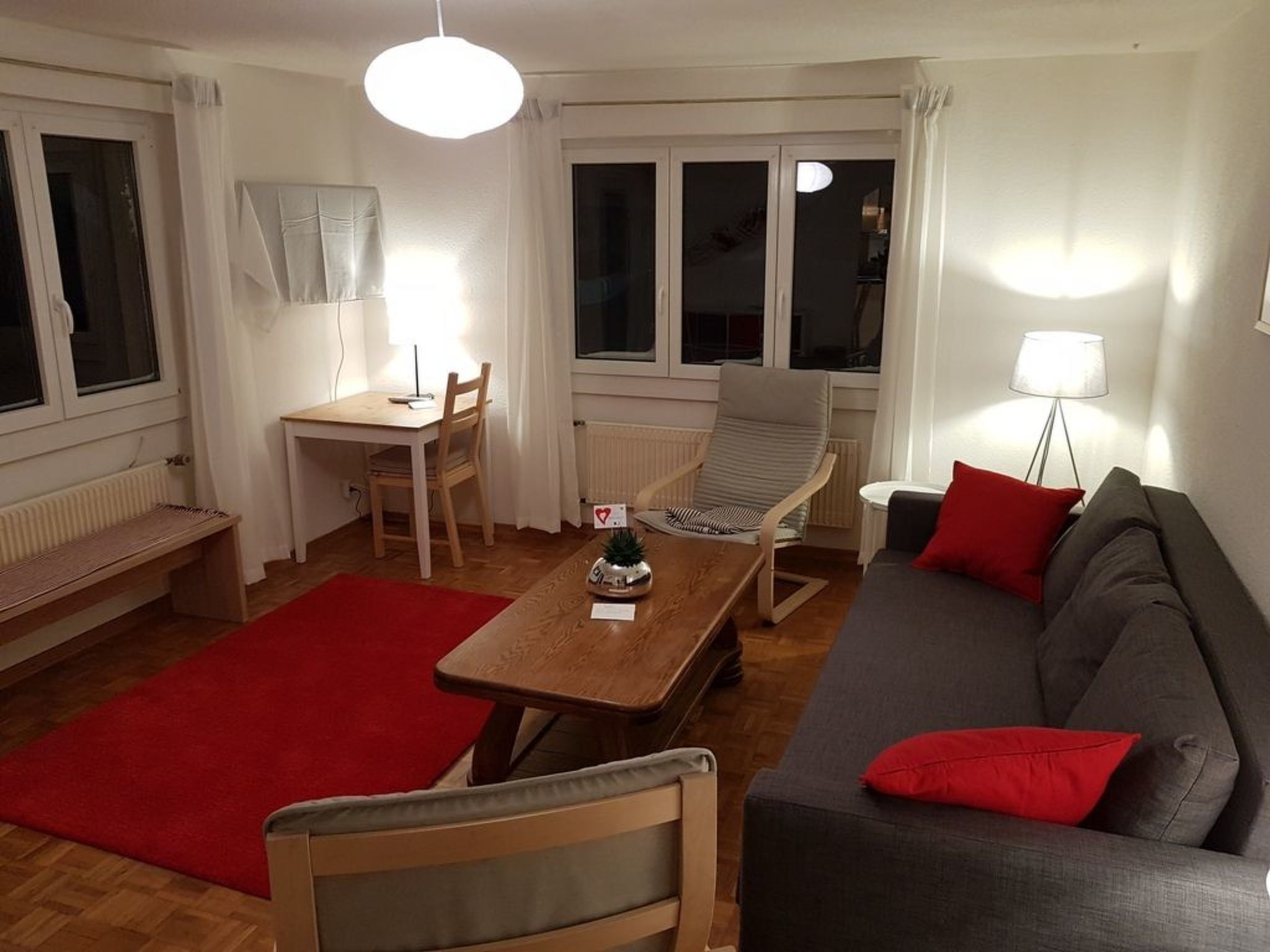 A living room with a sofa, a dining table, a chair, a red rug, and a lamp.