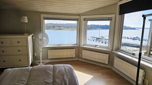 Camera da letto con un'ampia finestra a bovindo, termosifone, comò, ventilatore e vista su un lago e un molo.