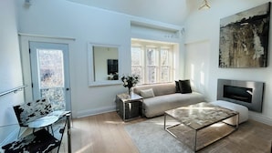 A modern living room with a sofa, coffee table, and a TV. There are windows, a door, and a painting on the wall. - Full Yorkville House 3Bed w/ Spa, Gym, Parking (Toronto)