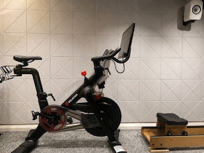 Een stilstaande hometrainer met een digitaal scherm, geplaatst in een ruimte met een betegelde muur en een klein houten constructie.
