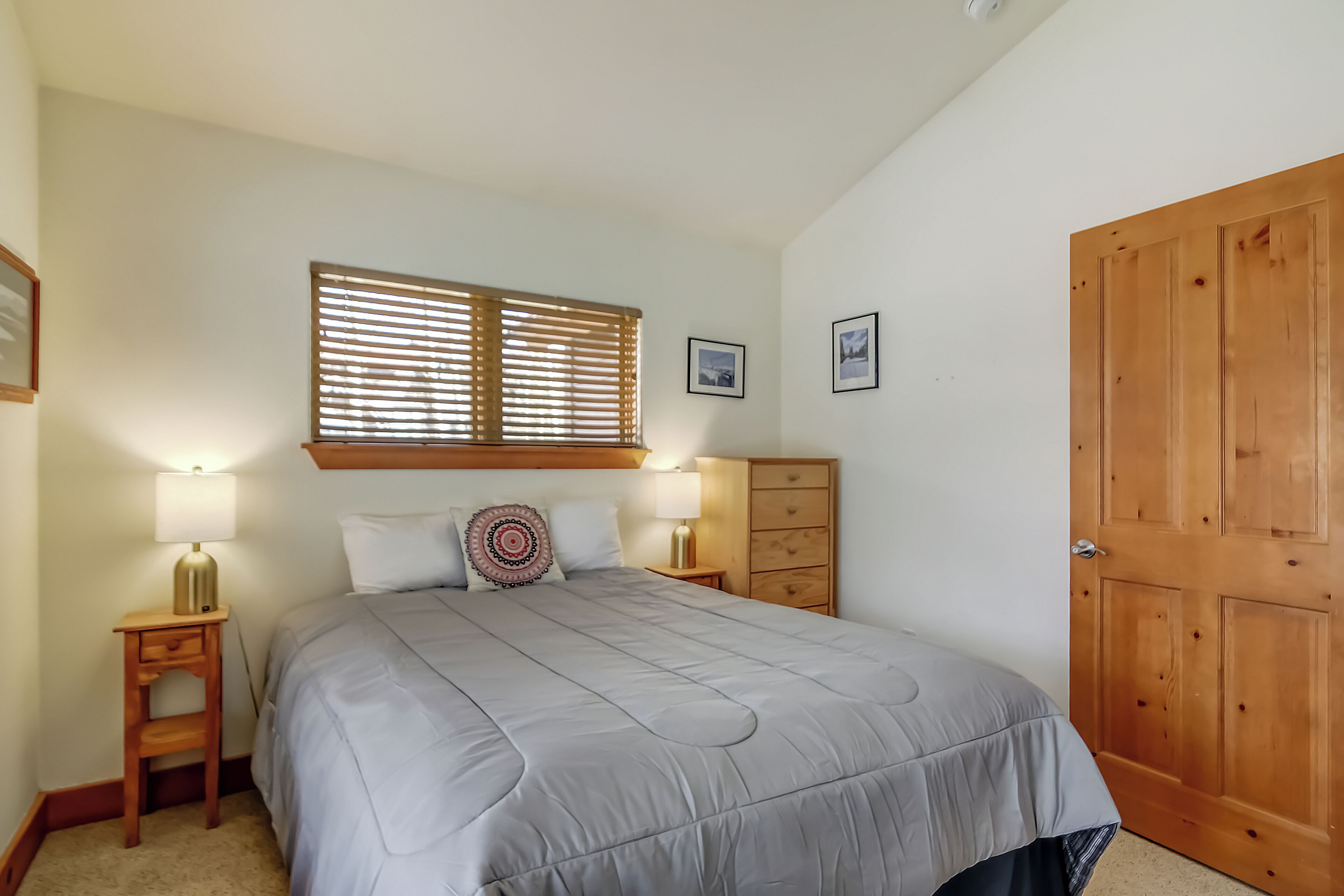 A bedroom with a bed, two bedside tables with lamps, a wooden wardrobe, and a window with blinds.