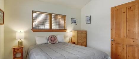 A bedroom with a bed, two bedside tables with lamps, a wooden wardrobe, and a window with blinds.