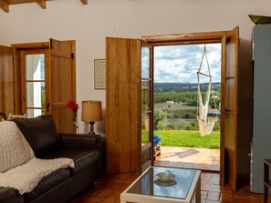 Ein gemütliches Wohnzimmer mit einem Ledersofa, einem Glastisch und Blick auf eine grüne Landschaft durch eine Holztür.