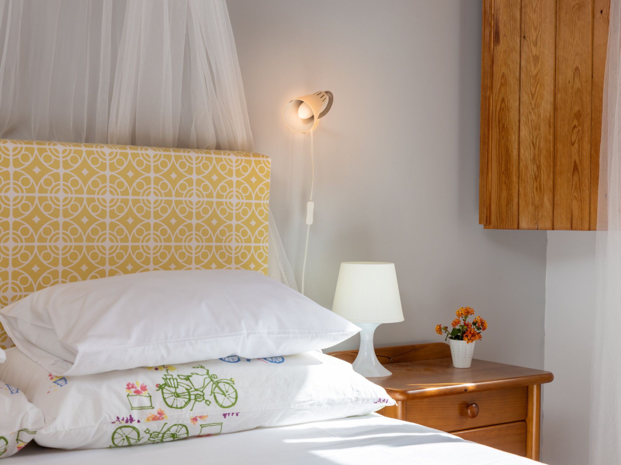 Un dormitorio con cama, mesita de noche con lámpara, mesita auxiliar con un florero y un armario de madera.