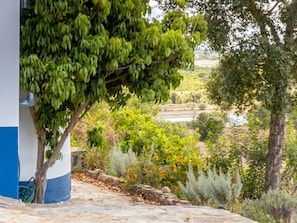 Ein steiniger Pfad führt zu einem Gebäude mit blauer und weißer Fassade, umgeben von üppiger Vegetation und mit Blick auf eine ferne Landschaft.