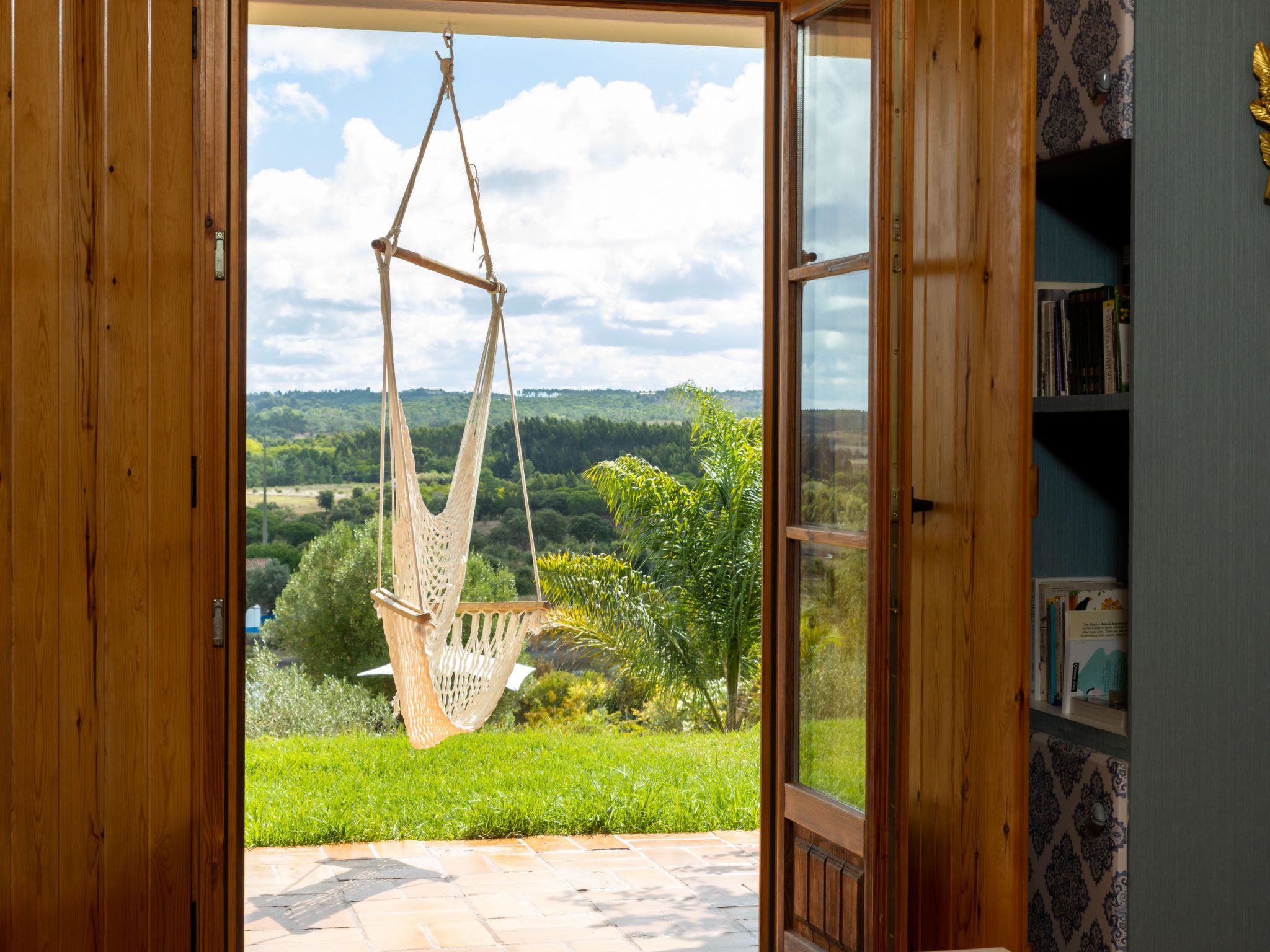 Puerta de madera abierta que da a una silla hamaca colgada con cuerdas, con vista a un paisaje verde y una estantería a la derecha.