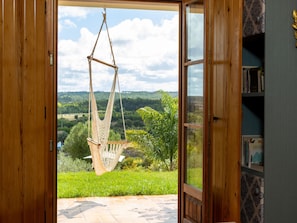 Eine offene Holztür, die zu einem Hängesessel führt, der an Seilen befestigt ist, mit Blick auf eine grüne Landschaft und einem Bücherregal zur rechten Seite.