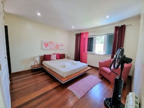 A bedroom with a bed, a bedside table, a chair, a window with curtains, and a standing fan. - VILLA FATI by Interhome (Tróia)