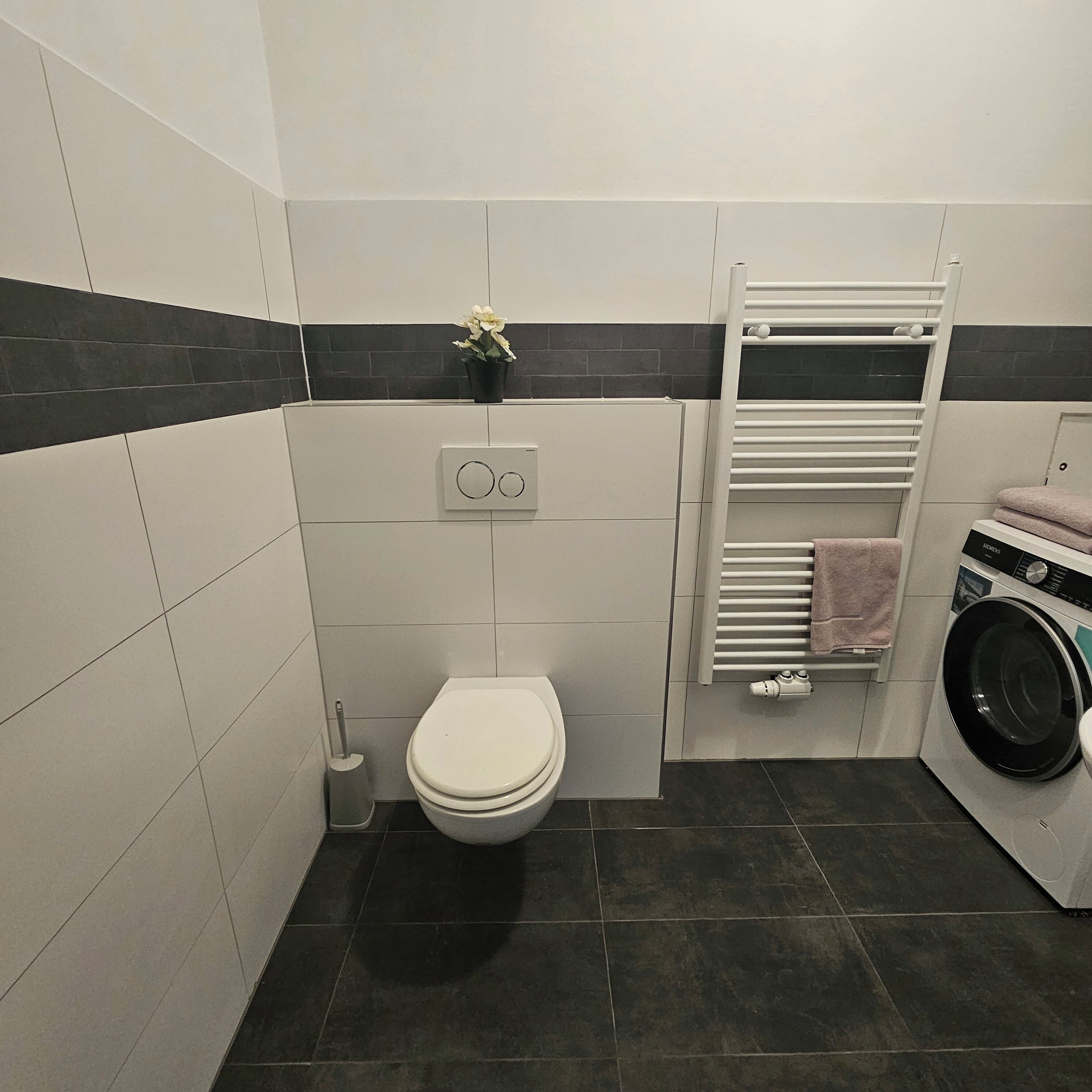 A compact bathroom with a toilet, a wall-mounted radiator, a washing machine, and a towel rack.