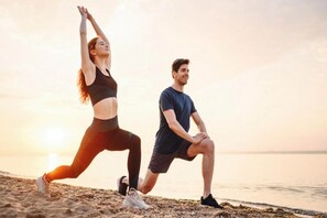 Two individuals performing stretching exercises on a beach with a sunset in the background.