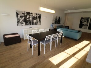 Un salon moderne avec un coin repas, comprenant une table en bois foncé et des chaises blanches, un canapé bleu et des œuvres d’art accrochées aux murs.