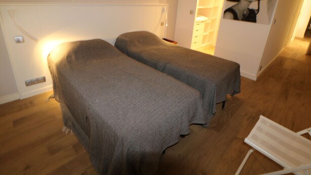 Ein Zimmer mit zwei Betten, die mit gemusterten Decken bezogen sind, einem weißen Stuhl und einem an der Wand befestigten Regal mit einem gerahmten Bild.