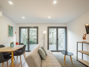 A modern living room with a sofa, dining table, chairs, and a large window with a view of a stone wall and greenery. - Apartment  by Interhome (Porto)