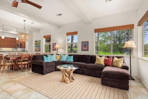 A spacious living area with a sectional sofa, dining table, and chairs, a coffee table, and large windows with a view of greenery. - NEW! Mauna Lani Beach Club Pass/BBQ on lanai/602 Fairways at Mauna Lani (Kamuela)