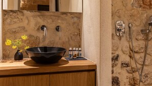 A bathroom with a stone wall, a wooden vanity, a black basin, a mirror, and a shower with a handheld showerhead. - Korona Boutique Hotel - Deluxe Room (Oitylo)