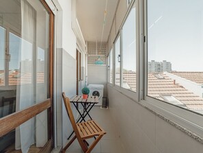 Un balcón con una silla de madera, una mesita con una planta en maceta, una lavadora y una lámpara colgante.