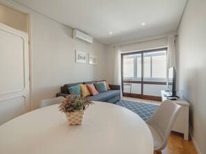 En moderne stue med et rundt bord, en sofa med mønstrede puder, et lille sofabord med en lampe og et vindue med gardiner.