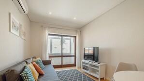 A modern living room with a grey sofa, colorful cushions, a flat-screen TV on a white stand, a patterned rug, and a large window with a view of buildings. - Apartment  by Interhome (Porto)