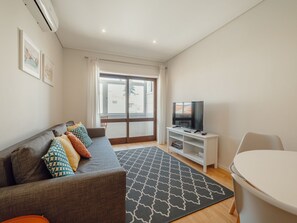 A modern living room with a grey sofa, colorful cushions, a flat-screen TV on a white stand, a patterned rug, and a large window with a view of buildings. - Apartment  by Interhome (Porto)
