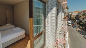 A balcony with a glass door, a bed with white linens, and a view of a street with parked cars. - Apartment  by Interhome (Porto)