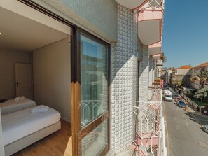 A balcony with a glass door, a bed with white linens, and a view of a street with parked cars. - Apartment  by Interhome (Porto)