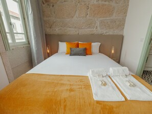 Ein Hotelzimmer mit Steinwand, einem Bett mit weißer und orangefarbener Bettwäsche, zwei Nachttischlampen und einer Handtuchgarnitur auf dem Bett.
