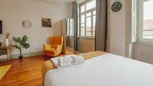 Une chambre d’hôtel avec un grand lit, une chaise, un bureau, une plante et une fenêtre avec des rideaux.