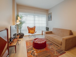 Un salon comprenant un canapé, un pouf rouge, une table basse en bois, un meuble pour la télévision et une plante en pot.