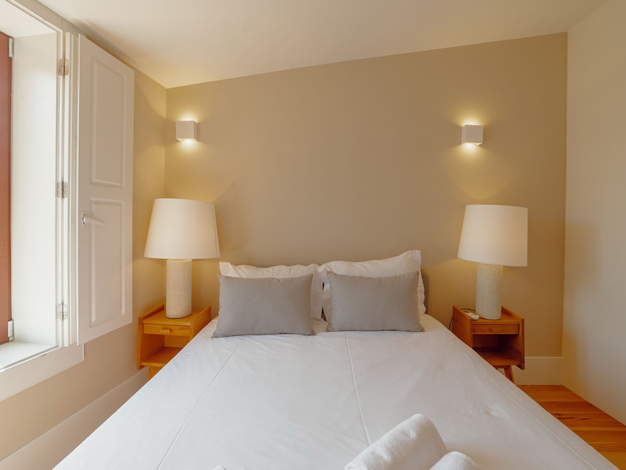A bedroom with a large bed, two bedside tables with lamps, a window with a door, and a wall-mounted light fixture.