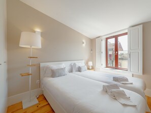 A hotel room with two beds, a standing lamp, and a window with a view of buildings. - Apartment  by Interhome (Porto)