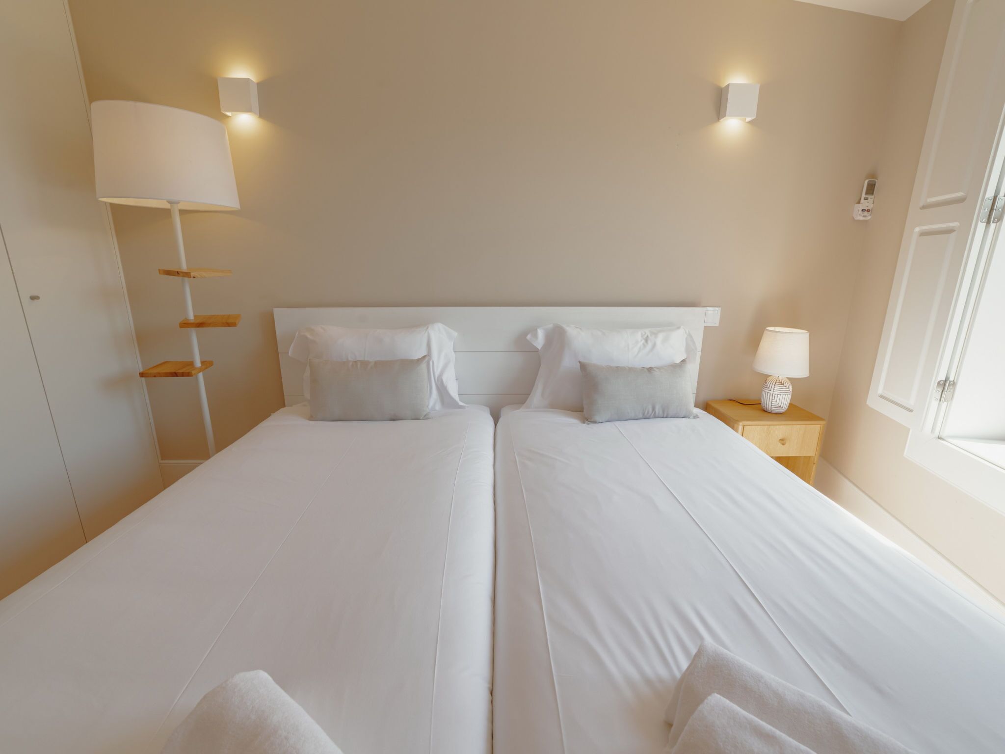 A double bed with white linens, a bedside table with a lamp, and a wall-mounted light fixture.