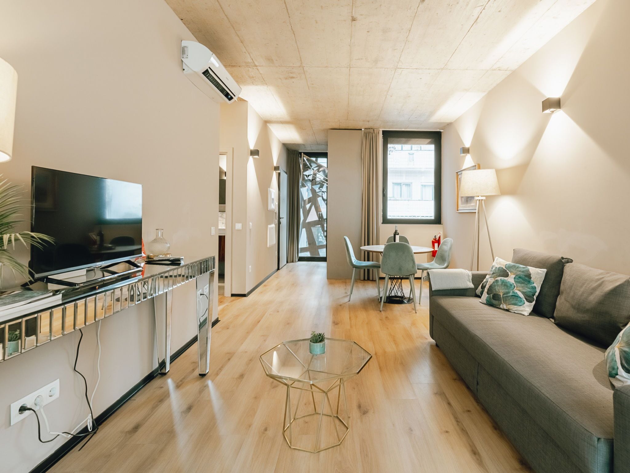 A modern living room with a flat-screen TV, a glass coffee table, a sofa with decorative pillows, and a dining area with a table and chairs.