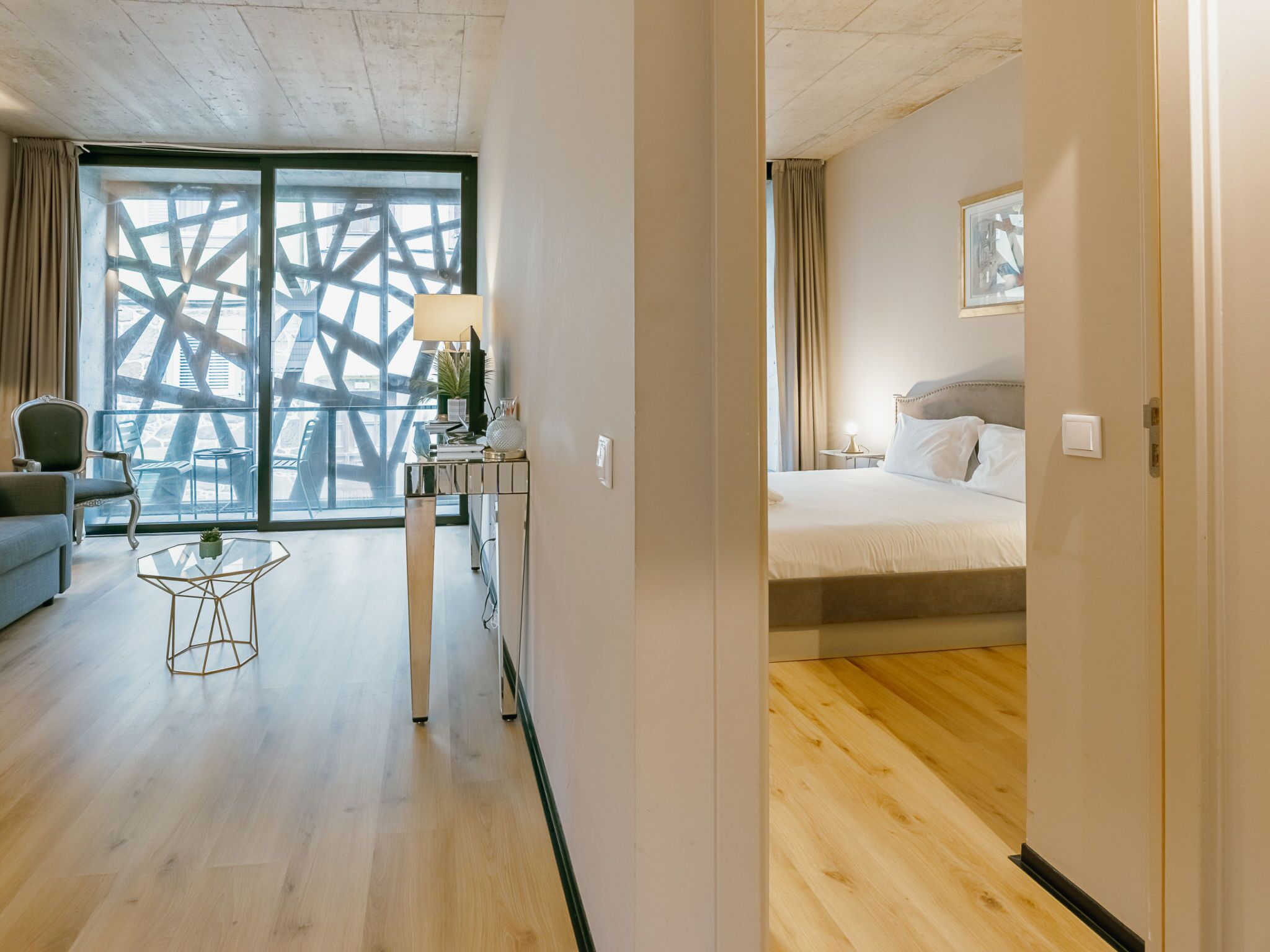 Ein Hotelzimmer mit Bett, Schreibtisch mit Lampe, Stuhl und Blick auf ein Gebäude durch ein großes Fenster.