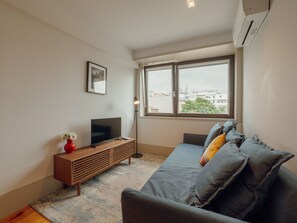 Un salon moderne avec un téléviseur à écran plat posé sur une console en bois, une grande fenêtre donnant sur la ville et un canapé gris agrémenté de coussins.