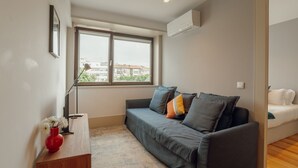 A modern living room with a grey sofa, a small wooden side table with a vase, and a window with a view of buildings. - Apartment  by Interhome (Porto)