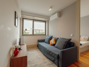 Uma sala de estar moderna com um sofá cinzento, uma pequena mesa de madeira com um vaso, e uma janela com vista para edifícios.
