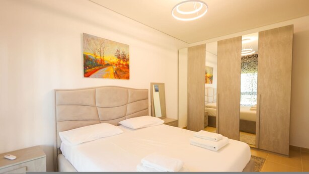 A modern bedroom with a large bed, a bedside table, a painting on the wall, and a mirror. - SanPietro Vacation Apartment 050 (Plazhi San Pietro)
